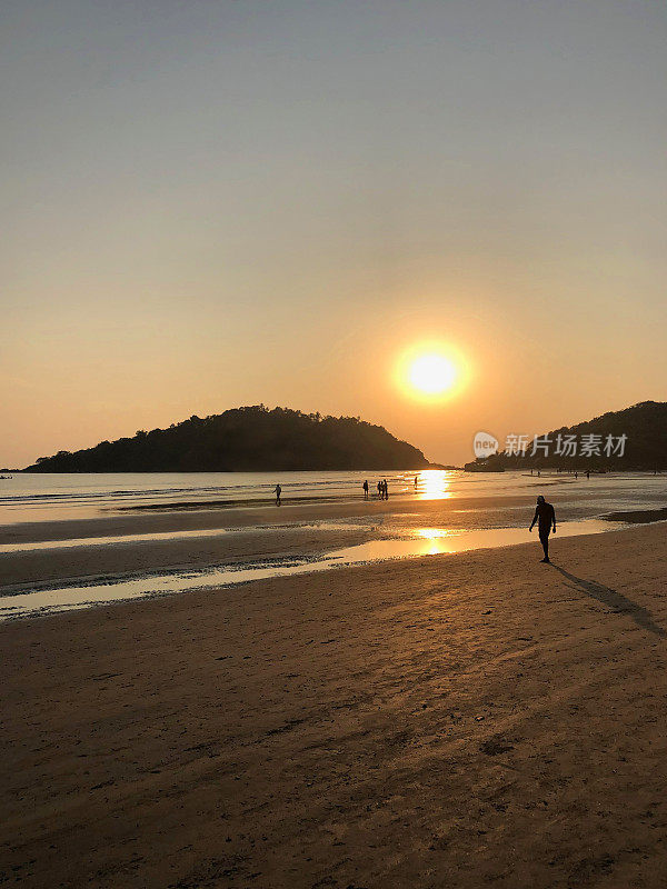 图片中，一名穿着短裤的男子走在印度果阿的帕洛伦海滩的海岸线上，夕阳下，海浪拍打着海滩