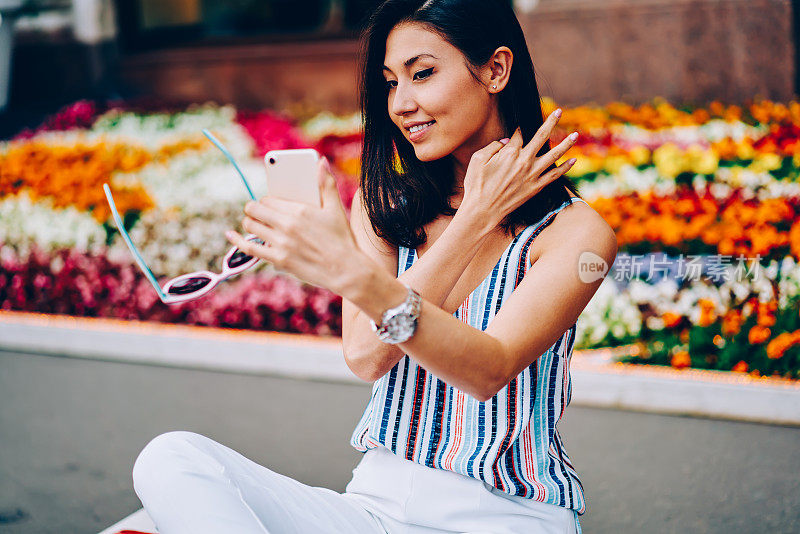 美丽的女性游客坐在广场上用智能手机拍摄照片，带着五颜六色的花，亚洲女性在漫游中使用智能手机和互联网为个人博客拍摄视频