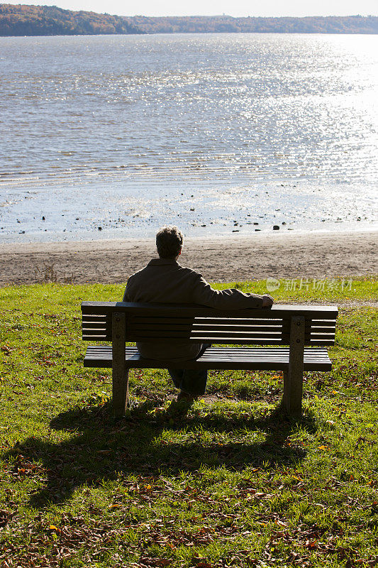 成熟的男人坐在长椅上俯瞰秋天的河流。
