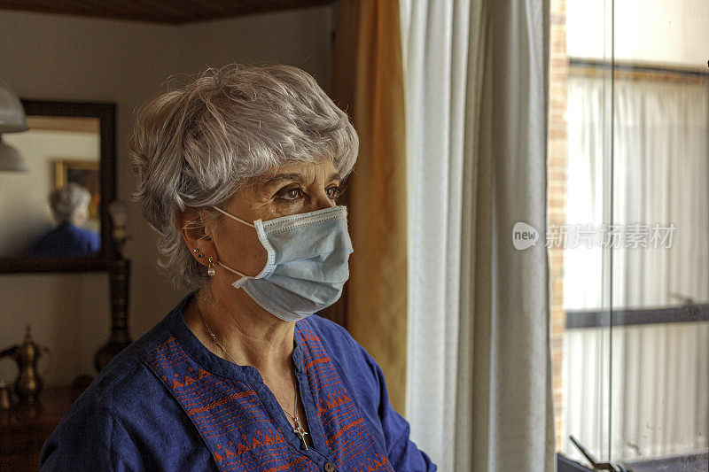哥伦比亚波哥大——在Covid-19疫情封锁的情况下，一位戴着口罩的拉丁美洲高级女士坐在家里靠窗的地方，外出购买杂货前查看天气