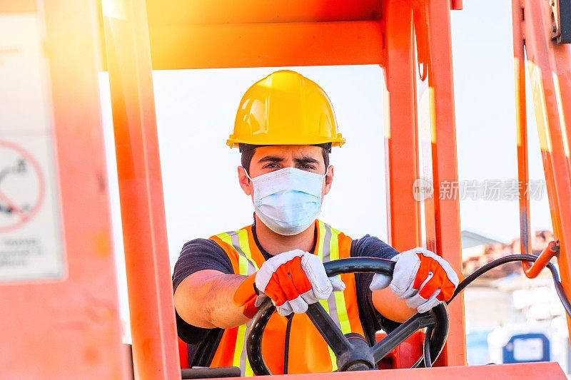 一名年轻的男性建筑工人戴着防护面罩看着摄像机，手里拿着一辆大型叉车的方向盘