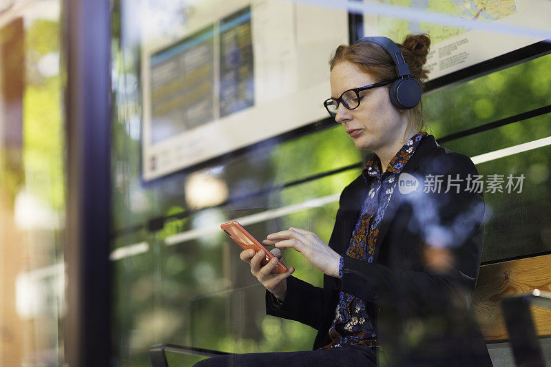一名女子在公交车站等车时使用手机和耳机