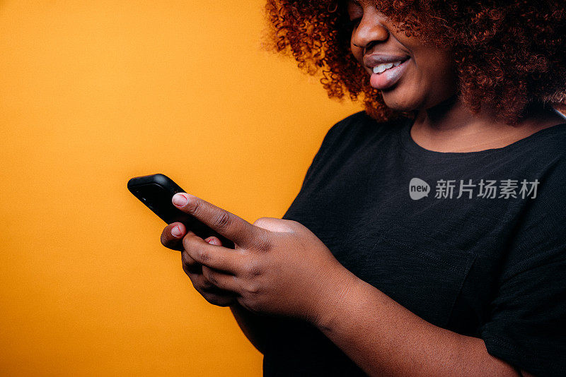 一个自信，美丽的年轻非洲裔美国妇女使用手持智能手机发送短信，网上银行，网上商店，或使用社交媒体在一个快乐的芥末黄色背景
