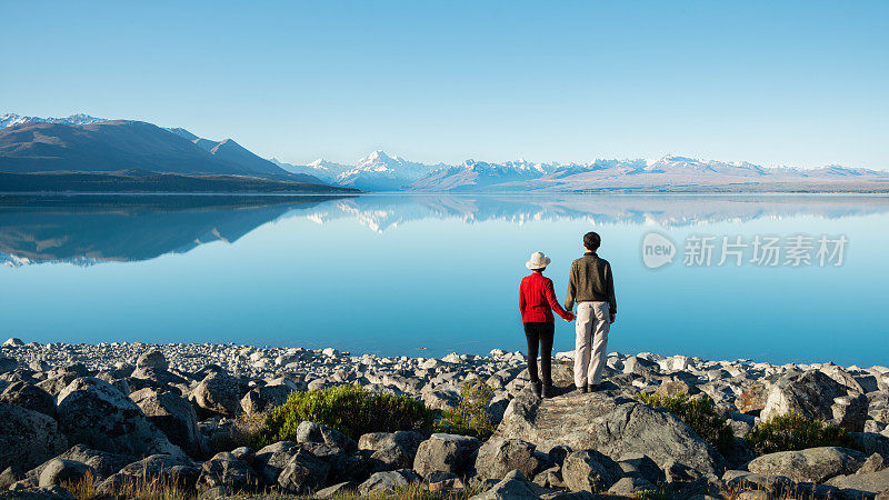 新西兰南岛，一对情侣站在普卡基湖岸边，手牵手，看着库克山倒映在清澈的湖水中