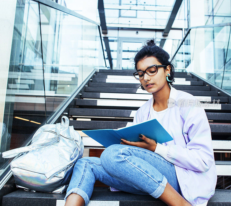 年轻可爱的印度女孩在大学大楼的楼梯上看书，戴着时髦的眼镜，生活方式的人的概念近距离