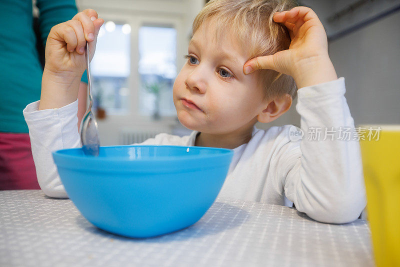 金发蹒跚学步的孩子坐在餐桌上，从碗里吃东西，看起来很累