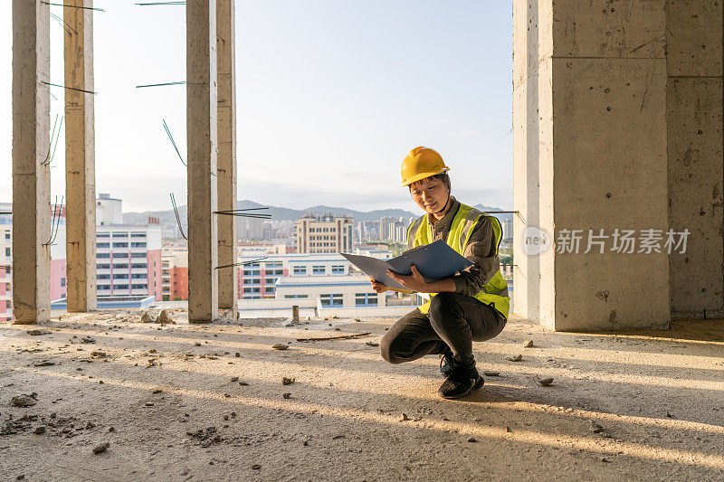 阳光透过建筑工地，一名女工程师蹲在地上拿着文件夹查看数据