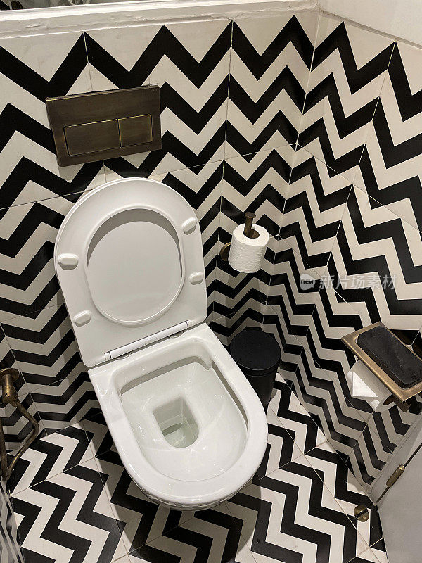 白色陶瓷浴室套房的特写图像，当代的壁挂式马桶与坐垫，单色之字形图案的墙壁和地砖，厕纸架上的厕纸卷，黑色脚踏箱，高架视图