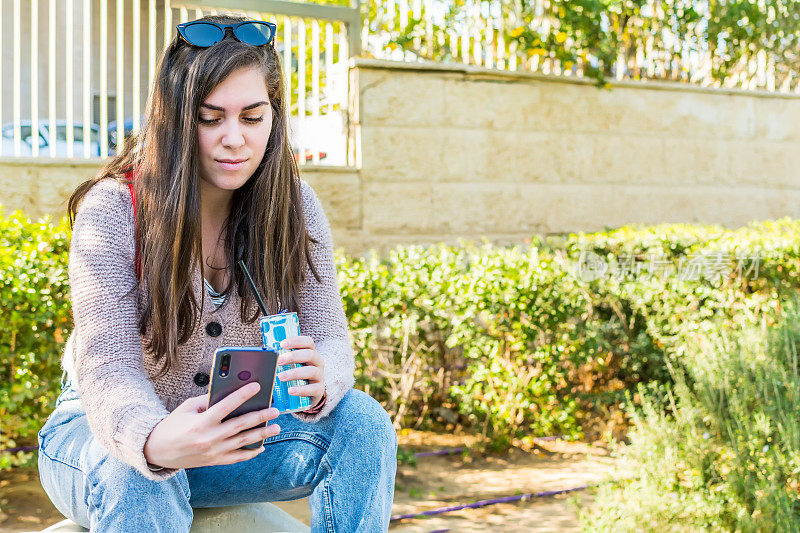 侧视图一个微笑的女性使用智能手机和苏打水坐在公园里