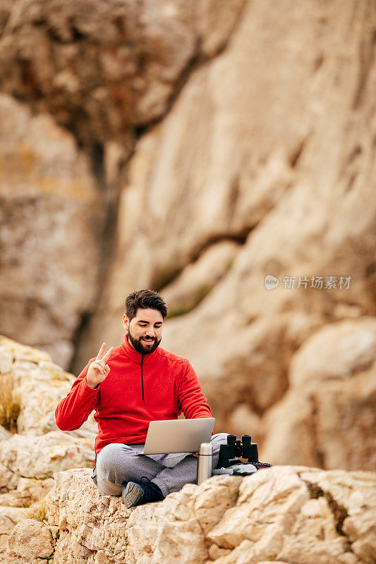 一个留着胡子的年轻徒步旅行者坐在岩石墙上，用两根手指指着他打开的笔记本电脑，背景是马略卡岛北部海岸的波伦萨湾