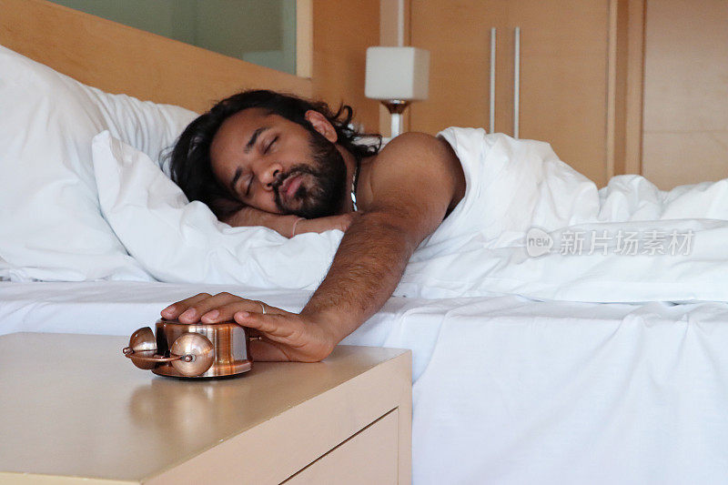 印度男子躺在酒店的双人床下被褥的形象，手伸向床头柜关闭打翻了青铜金属盒复古闹钟，双铃脸朝下躺着，专注于前景