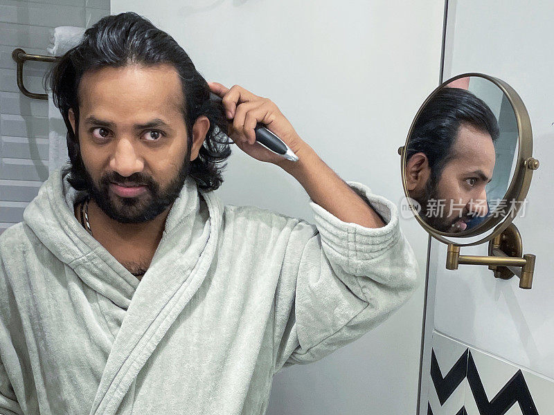 印度男人穿着浴袍站在浴室里，用梳子梳理凌乱潮湿的头发，剃须镜里的倒影，自我护理和修饰的概念