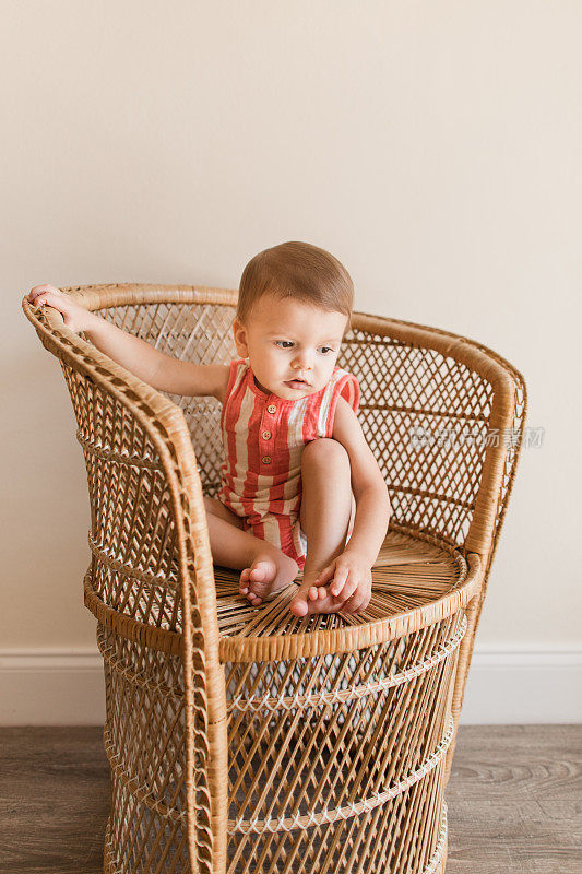 2023年，南佛罗里达，一个可爱快乐的17个月大的小男孩，浅棕色的头发和棕色的眼睛，穿着红色和奶油色的条纹纱布连衣裤，赤脚坐在一张老式藤椅上。男婴有轴后多趾畸形