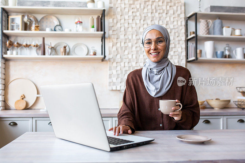 戴着头巾的年轻穆斯林妇女在家里的厨房里吃饭。喝咖啡，用笔记本电脑，休息一下，看着相机，微笑