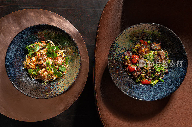 小牛肉、蘑菇和蔬菜荞麦面和海鲜蛋面放在黑色的陶瓷盘子里，放在铜桌子和皮椅子上。