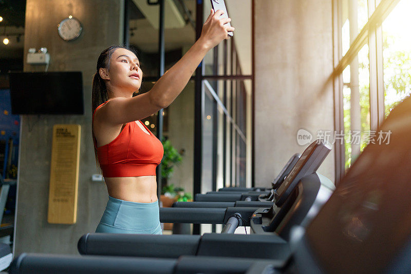 在室内健身俱乐部，一名亚洲女性在跑步机上休息，用智能手机在灿烂的阳光下自拍