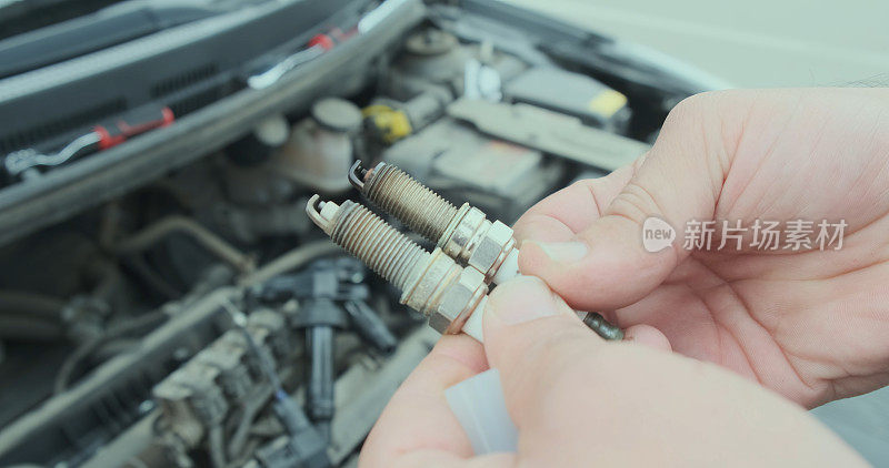 汽车修理工手里拿着两根汽车蜡烛，一根是干净的，另一根是浓重的黑烟。背景中凸起的汽车引擎盖。替换蜡烛的概念。