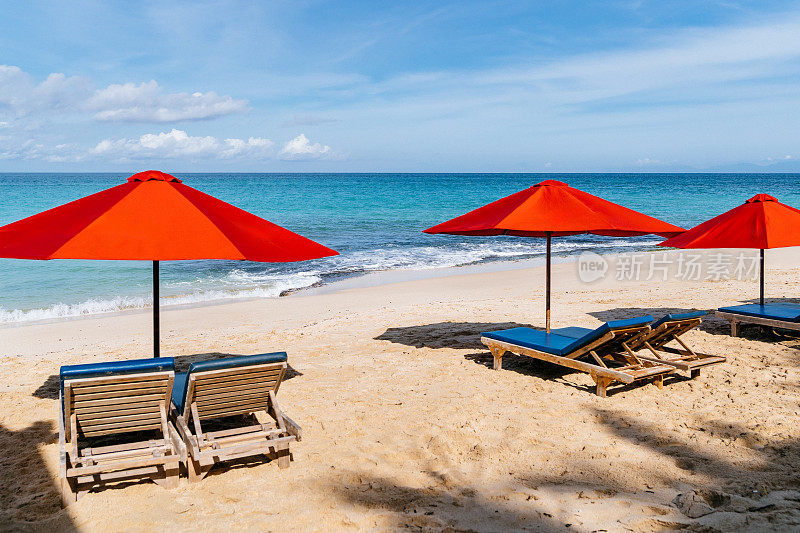巴厘岛，阳光海滩逃生:充满活力的红色雨伞，金色沙滩上的蓝色躺椅，清澈的海水和蔚蓝的天空。
