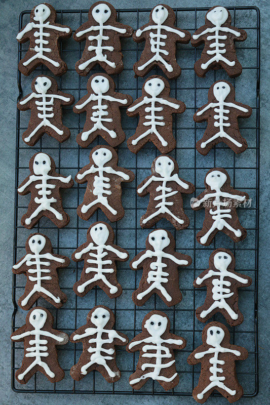 全框图片:一排排自制的人形巧克力饼干放在烤架上，上面是防油的羊皮纸烘焙纸，饼干用白色糖霜裹成骷髅状，居高临下，万圣节主题的派对食品