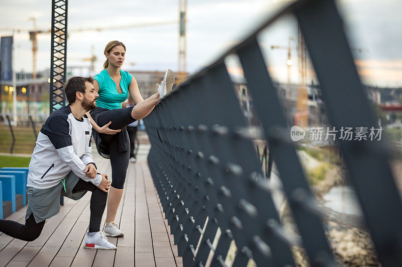 现代一对在大桥上跑步的夫妇正在跑步后伸展身体。英俊的运动员和他美丽的女友正在城市大桥上慢跑热身。