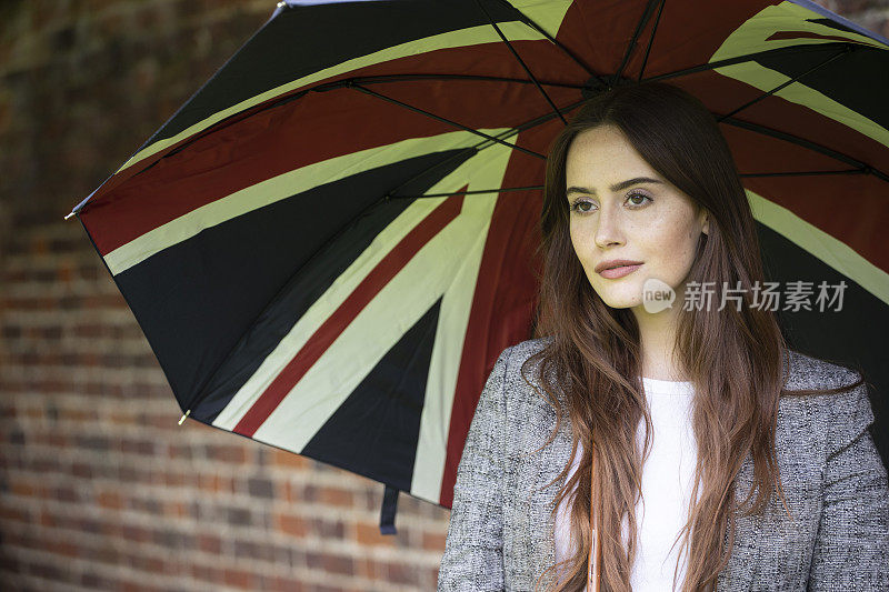 一位拿着英国国旗伞的年轻女子，站在一堵砖墙旁。她穿着一件漂亮的灰色定制夹克。
