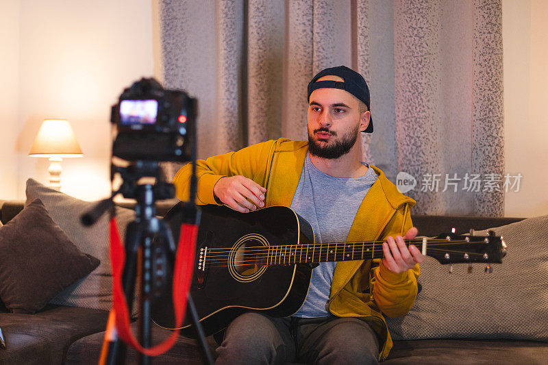 一个留着大胡子的年轻人一边在家里弹吉他，一边用单反相机为YouTube录制教程