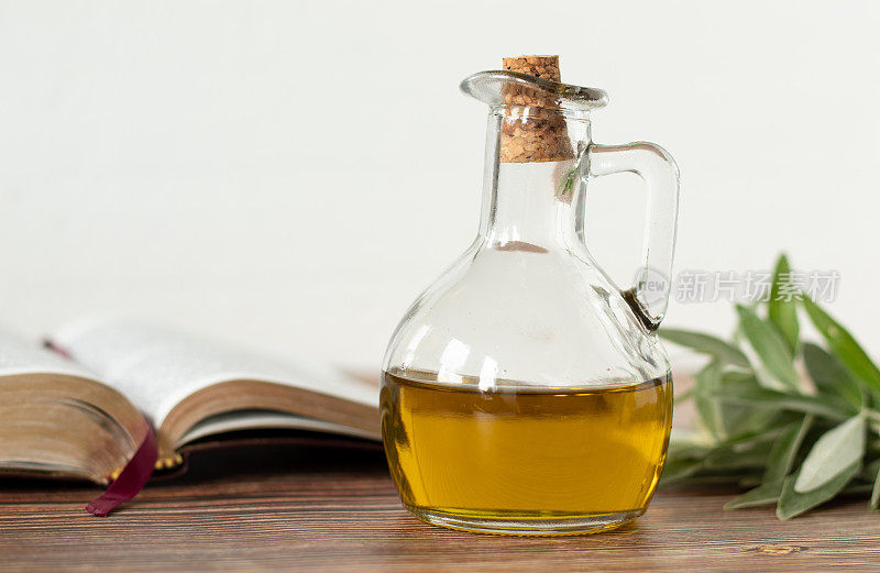 纯橄榄油装在玻璃罐里，一本打开的圣经，白色背景的木桌上放着一根绿色的橄榄枝