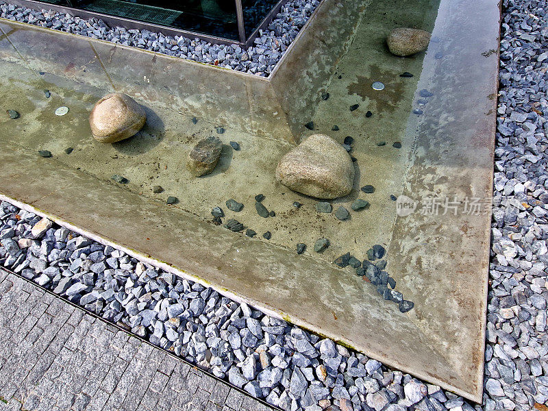 圆形的巨石从河里捞出来，放进一个有氯化水的池塘里。巨石露出水面的地方有一层盐壳。混凝土，水池，砾石人行道，角落，细节