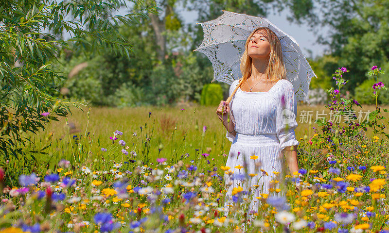 年轻的金发女子穿着白色的裙子，拿着白色的雨伞站在阳光下盛开的花坛