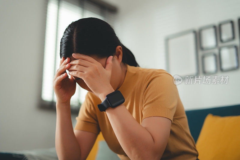 一名亚洲妇女正坐在家里客厅的沙发上饱受头痛之苦。