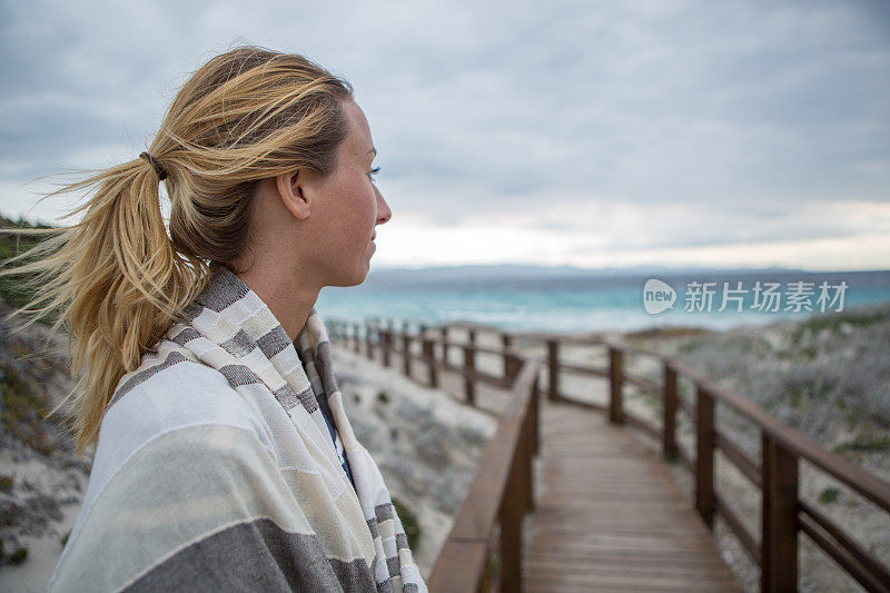 年轻女子裹着毯子走在木制的海滩小路上