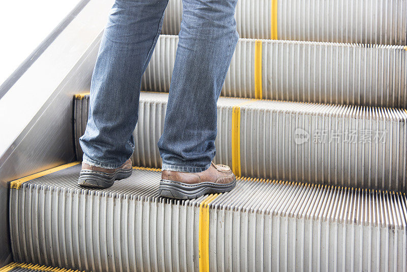 男士穿牛仔裤、皮鞋。站在自动扶梯上