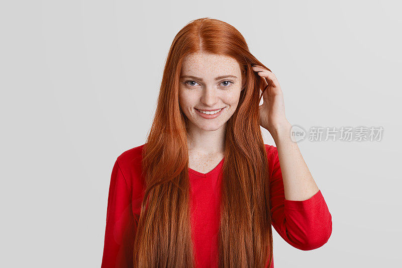肖像的红头发年轻女性与长头发，有雀斑的脸，愉快的微笑，触摸头发，孤立在白色的背景。姜女在工作室摆姿势，高兴被拍照。
