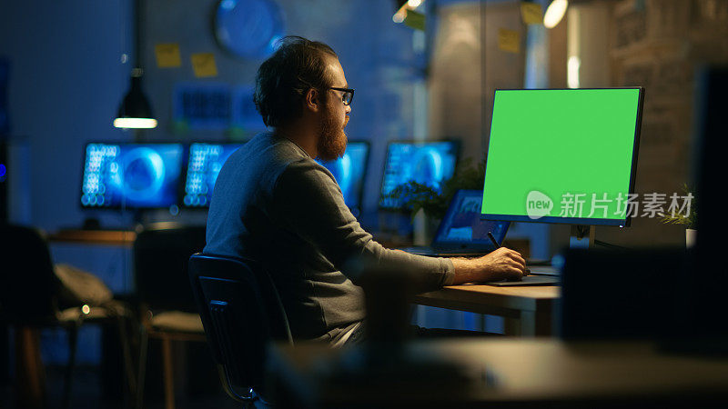 有创意的大胡子男人在一台个人电脑上工作，电脑上有独立的绿屏模型。他深夜独自在办公室工作。