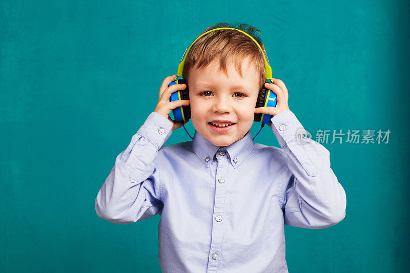 帅气的小男孩戴着明亮的耳机站着，通过智能手机听着音乐，靠在绿色的蓝色墙壁上