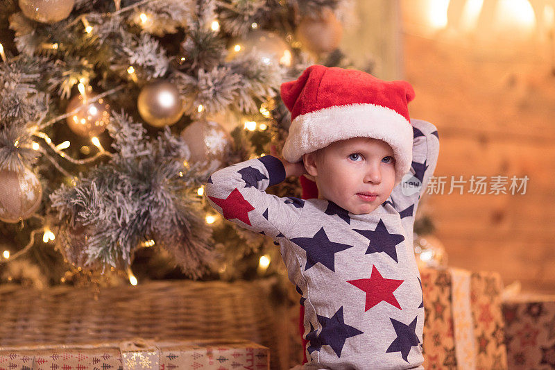 戴着圣诞老人帽子的快乐微笑的小男孩等待除夕打开礼物。圣诞节的概念与欢笑的孩子在圣诞树装饰花环和球