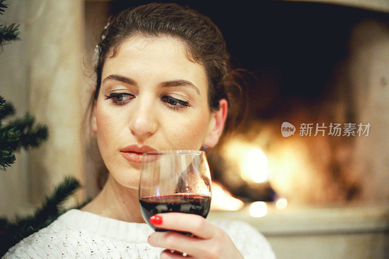 舒适的家。近距离的年轻女子喝红葡萄酒靠近壁炉。