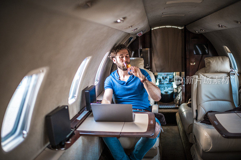 年轻孤独的富人坐在私人飞机上用笔记本电脑喝着酒