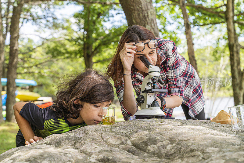 一个十几岁的女孩，一个17岁的妹妹教她9岁的弟弟如何使用显微镜，在阳光明媚的夏日，在湖边户外学习大自然。