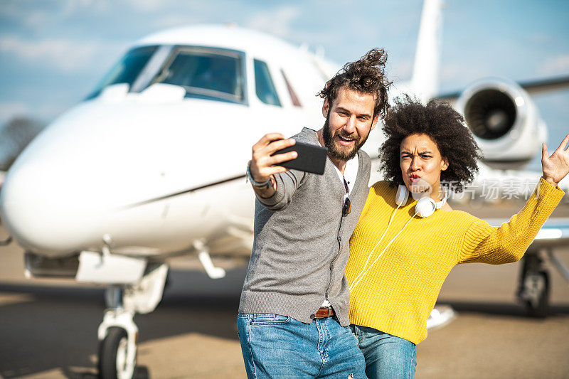 富有的非洲裔美国女孩挥手，与她的伴侣在一架停在机场跑道上的私人飞机旁自拍