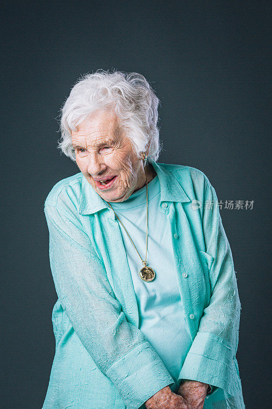 甜美，快乐，快乐，聪明的老年白人妇女微笑着，穿着她最喜欢的蓝绿色衬衫，站着向下看和欣赏在工作室与复制空间的灰色背景的东西