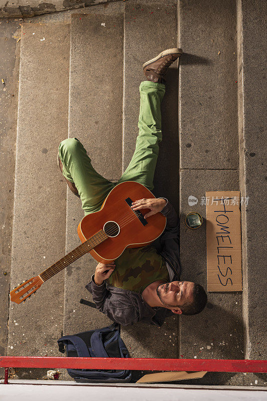一名因酗酒而失去意识的流浪汉躺在楼梯上，手里拿着他的吉他和盒子等待捐赠