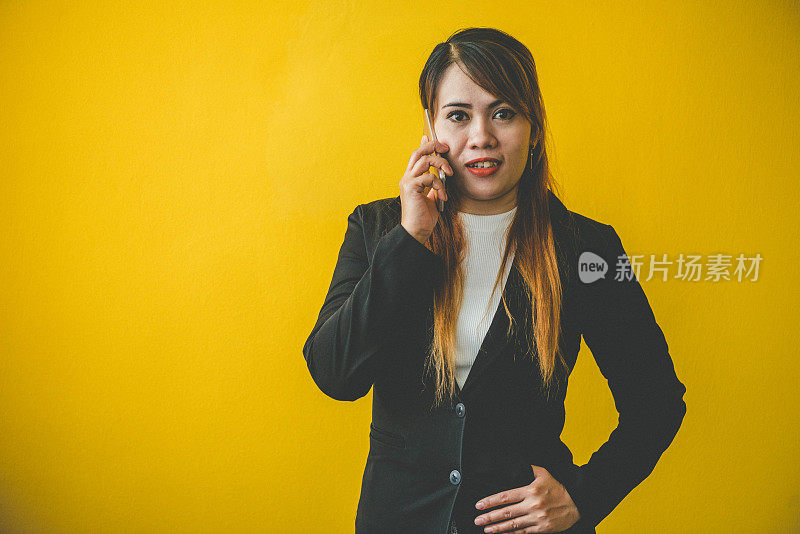 亚洲女人在黑色西装是快乐的，拿着手机在黄色的背景