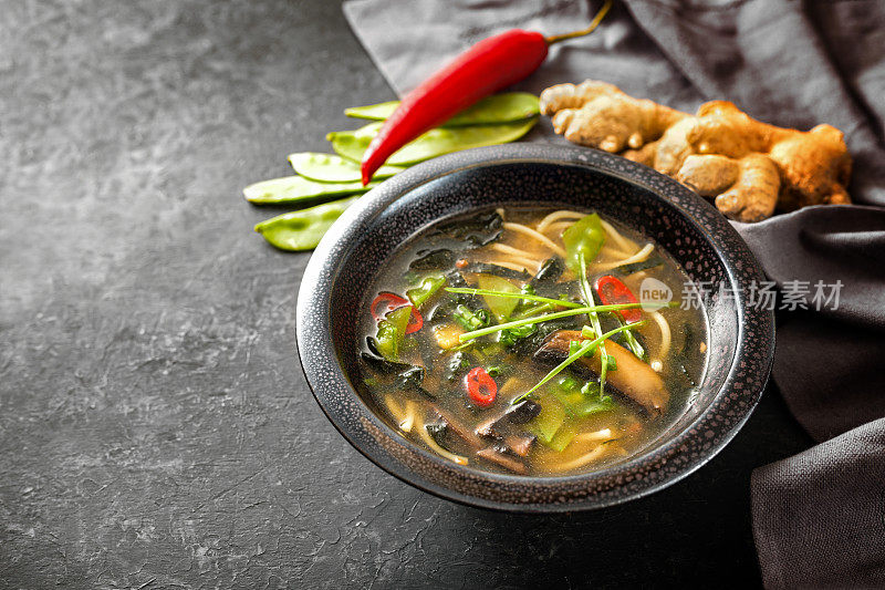亚洲蔬菜汤，有面条、蘑菇、糖豆、海苔、生姜和辣椒，传统上用味增酱调味，放在黑色陶瓷碗里，背景是深色的，复制空间，选择重点