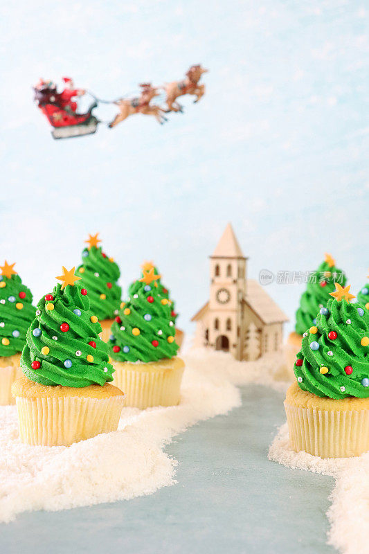 雪景的圣诞形象，一批自制的，圣诞树设计的纸杯蛋糕排列在人行道上，圣诞老人雪橇和驯鹿飞行，木制教堂模型，糖霜雪，重点在前景