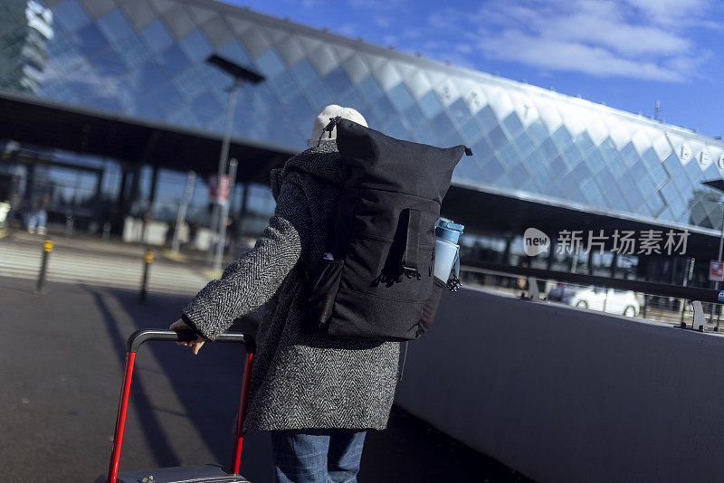 一名女游客拖着行李箱，背着登山背包走向机场的后视图