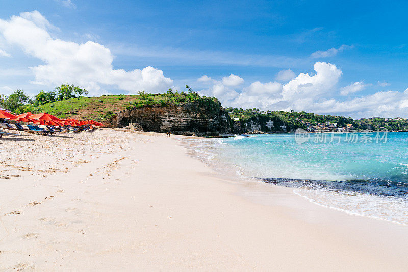 阳光明媚的一天在热带海滩:白色的沙滩，清澈的蓝色海水，红色的雨伞，和悬崖边的绿色植物在巴厘岛。