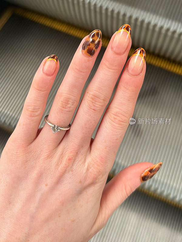 在自动扶梯上无法辨认的女性特写，左手展示订婚戒指和指甲艺术，展示豹纹图案指甲油美甲设计，移动的楼梯台阶背景，高架视图