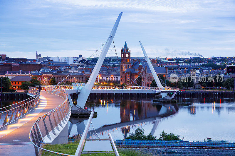 德里,爱尔兰。北爱尔兰文化之城伦敦德里的和平桥被灯光照亮，背景是市中心。夜晚多云的天空映着黄昏的河水