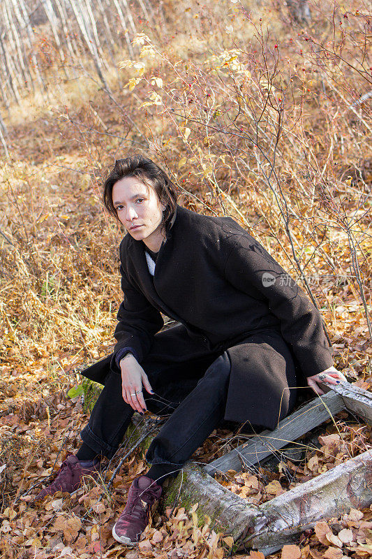 在宁静的秋林里，一位穿着黑色外套的年轻女子坐在一根圆木上。